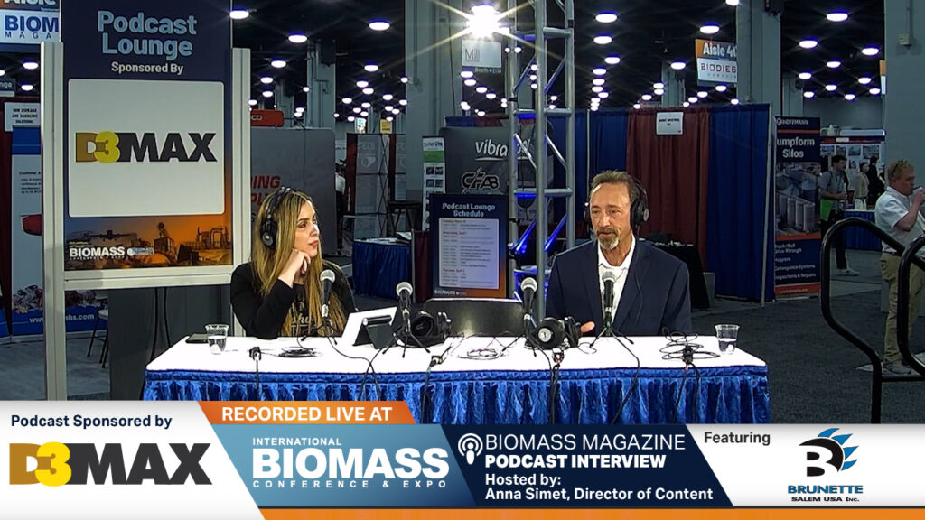 Image shows a woman with long hair and a man dressed in a suit sitting at a table with headphones on. Each person has a microphone in front of them. They are conducting a live interview at a trade show conference